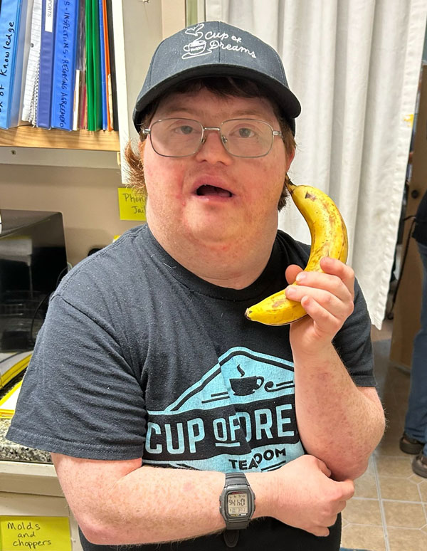Joshua smiling in a gray cap and Cup of Dreams Tearoom T-shirt, holding up a banana.