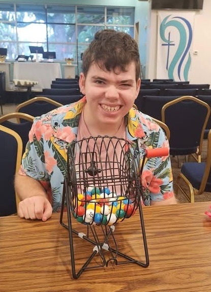 Sean smiling in a floral shirt beside a bingo cage at a SociAbilities game night.