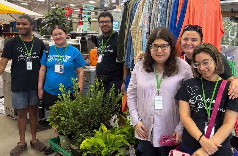 Six PossAbilities Plus participants in Cup of Dreams shirts smiling and holding plants on a store outing.