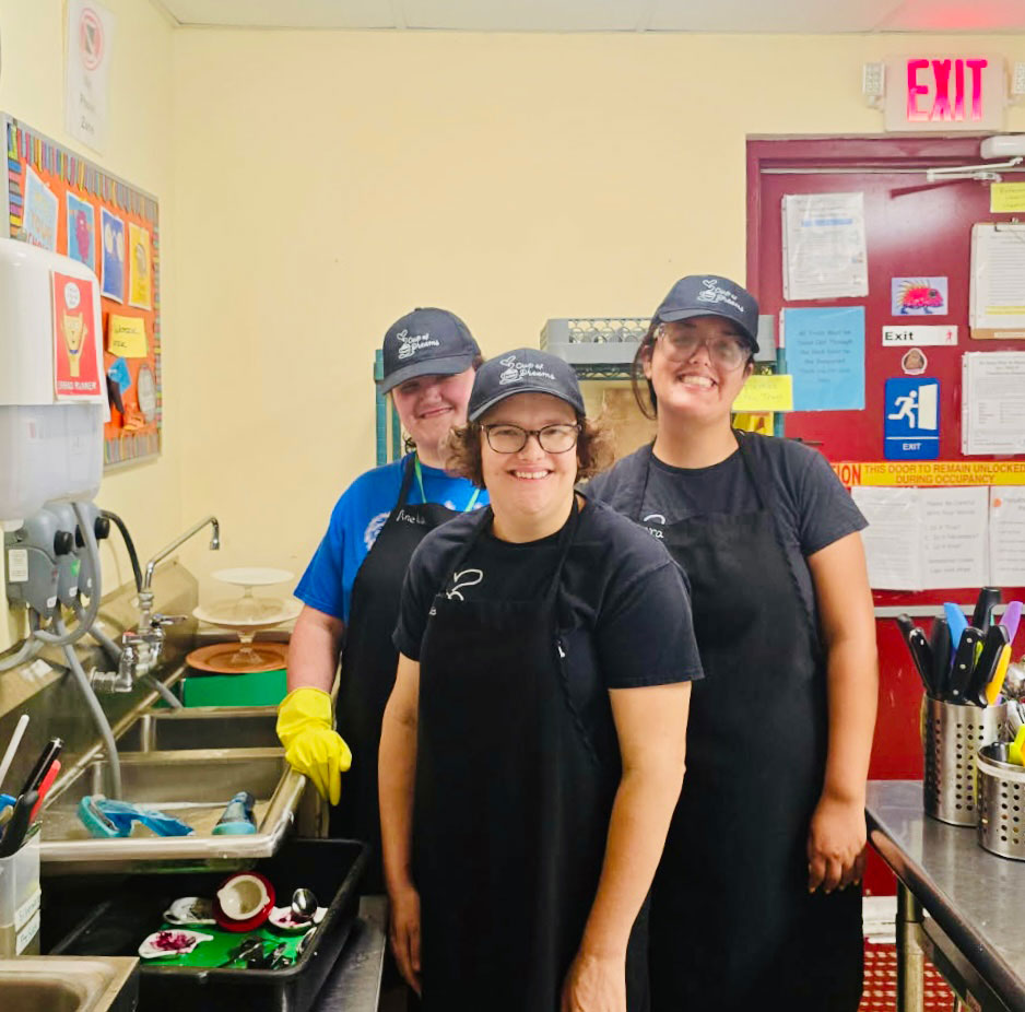 Three participants in caps and black aprons smiling in the Cup of Dreams training kitchen.