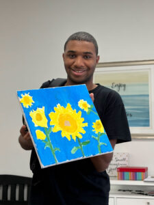 Conner smiling and holding up his painting of yellow sunflowers on a blue background.