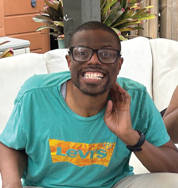 Frank smiling in glasses and a teal T-shirt, resting one hand by his face.