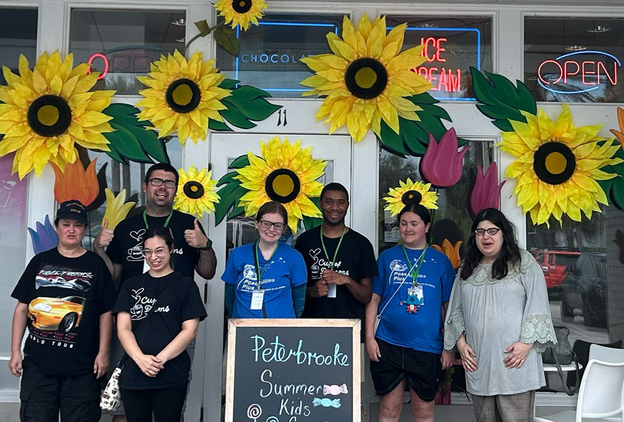 Eight PossAbilities Plus participants and staff smiling together outside Peterbrooke Chocolatier, its windows decorated with giant paper sunflowers.