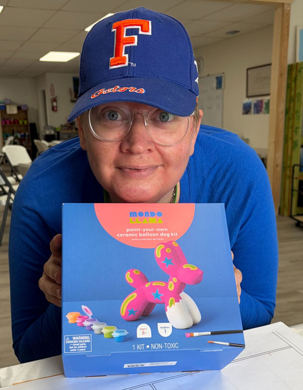 Vanessa smiling in glasses and a Florida Gators cap, holding up a paint-your-own ceramic balloon dog kit.