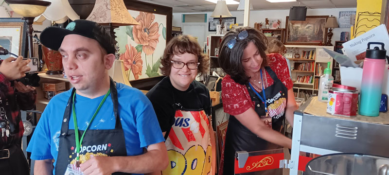 Three participants in aprons working the popcorn station on the Unique Boutique sales floor, smiling as they serve customers.