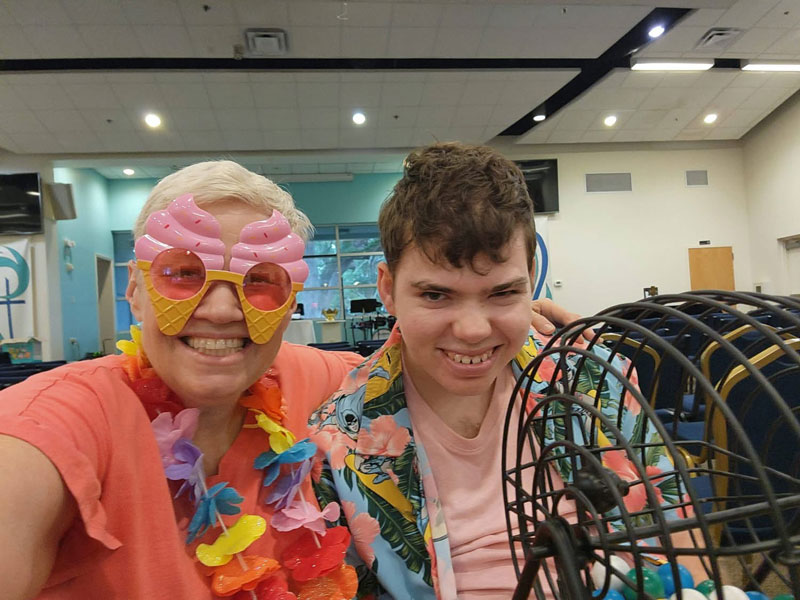 A participant and a volunteer in leis and party glasses smiling beside a bingo cage at a SociAbilities game night.